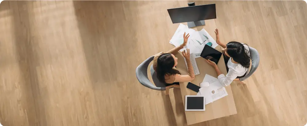 Overhead view of two women sitting at a desk, discussing work with documents, a computer monitor, and a tablet on the table