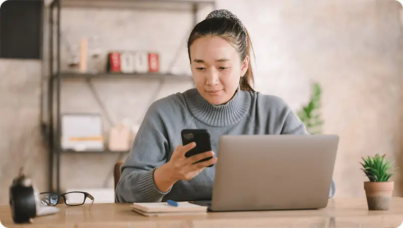 Asian female business person checks smart phone whilst worknig on laptop.